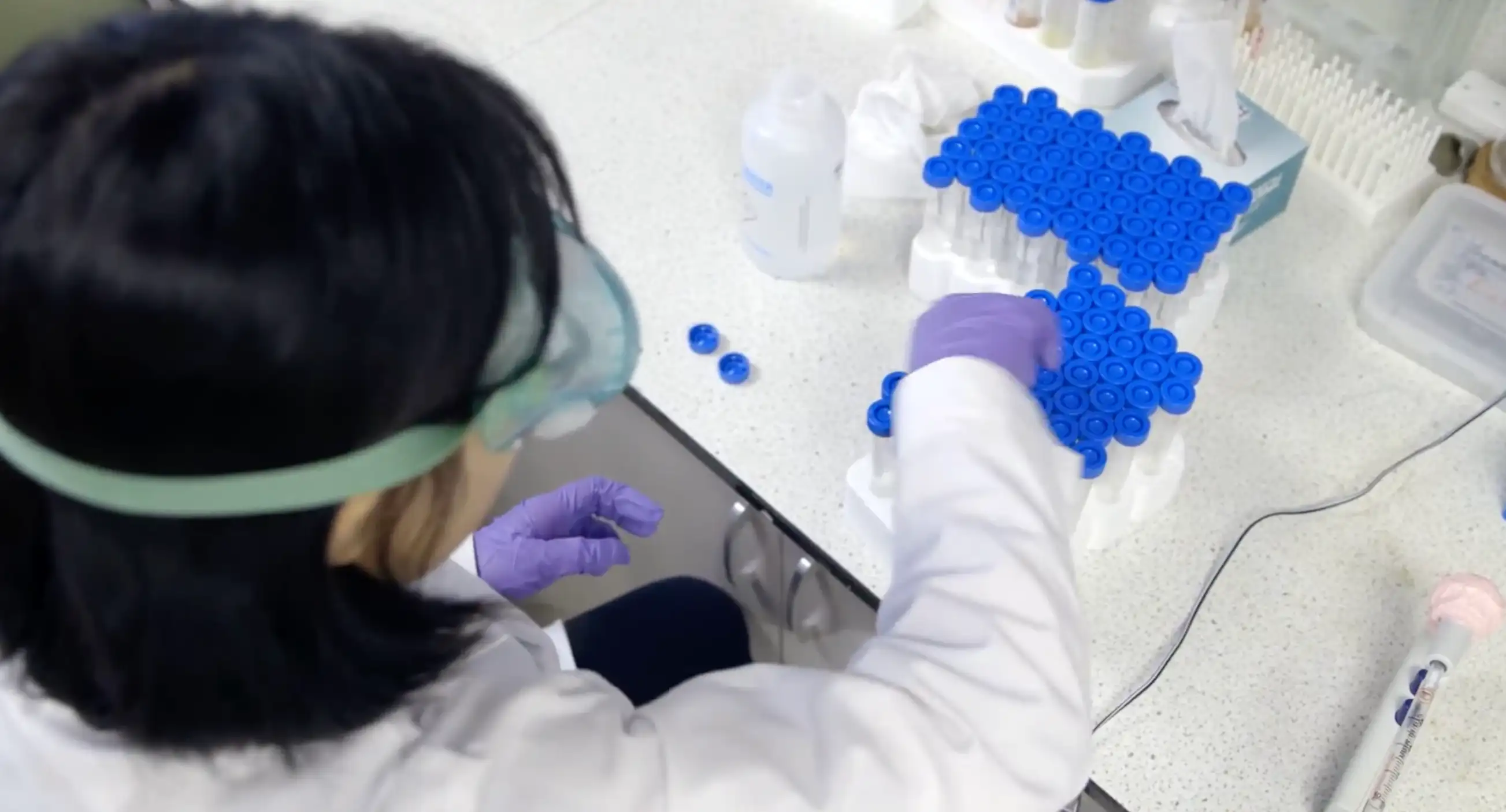 A researcher in a lab coat and safety goggles working with racks of blue-capped tubes